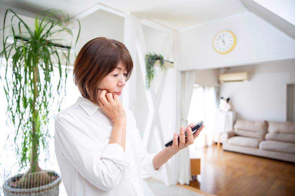 自宅でスマホを見ながら考え込む女性のイメージ、一人で情報を集めて悩む様子を表す写真